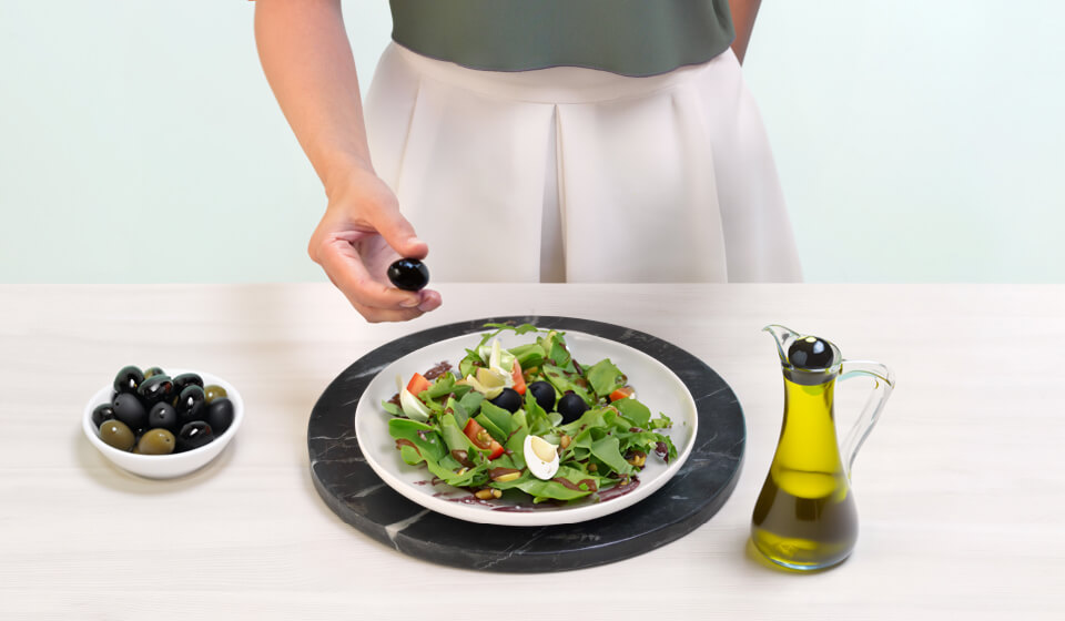 Eine Person gibt eine Olive auf einen Salatteller neben einer Ölkanne und einer Schale Oliven.