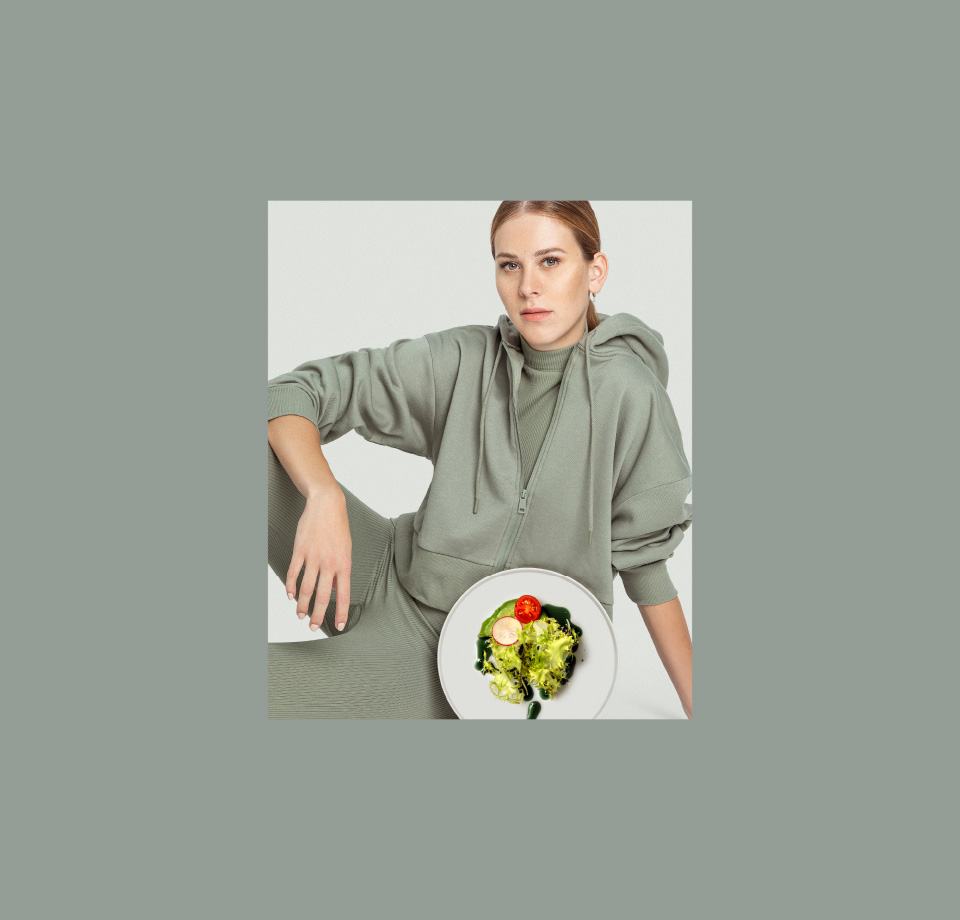 Eine Frau in einem grünen Hoodie sitzt mit einem Teller Salat vor sich , der mit original steirischem Pelzmann Kürbiskernöl garniert ist. Der Hintergrund ist neutral.