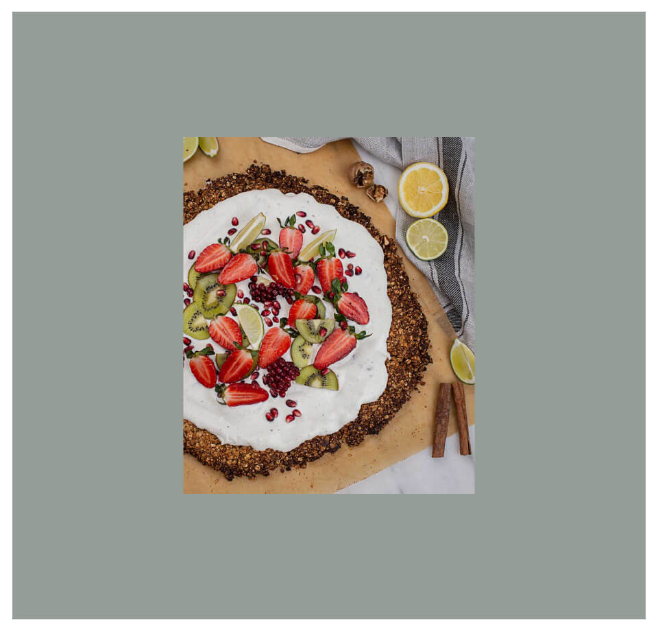 Pelzmann Granola Pizza mit Früchten und weißen Belag, umgeben von Zimtstangen, Limettenscheiben und Granatapfelkernen auf einer Unterlage.